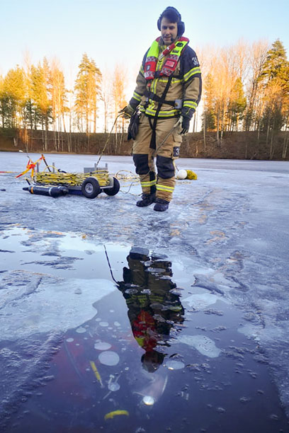 Brandman står på isen vid en vak med flytväst och en liten maskin på hjul med handtag.