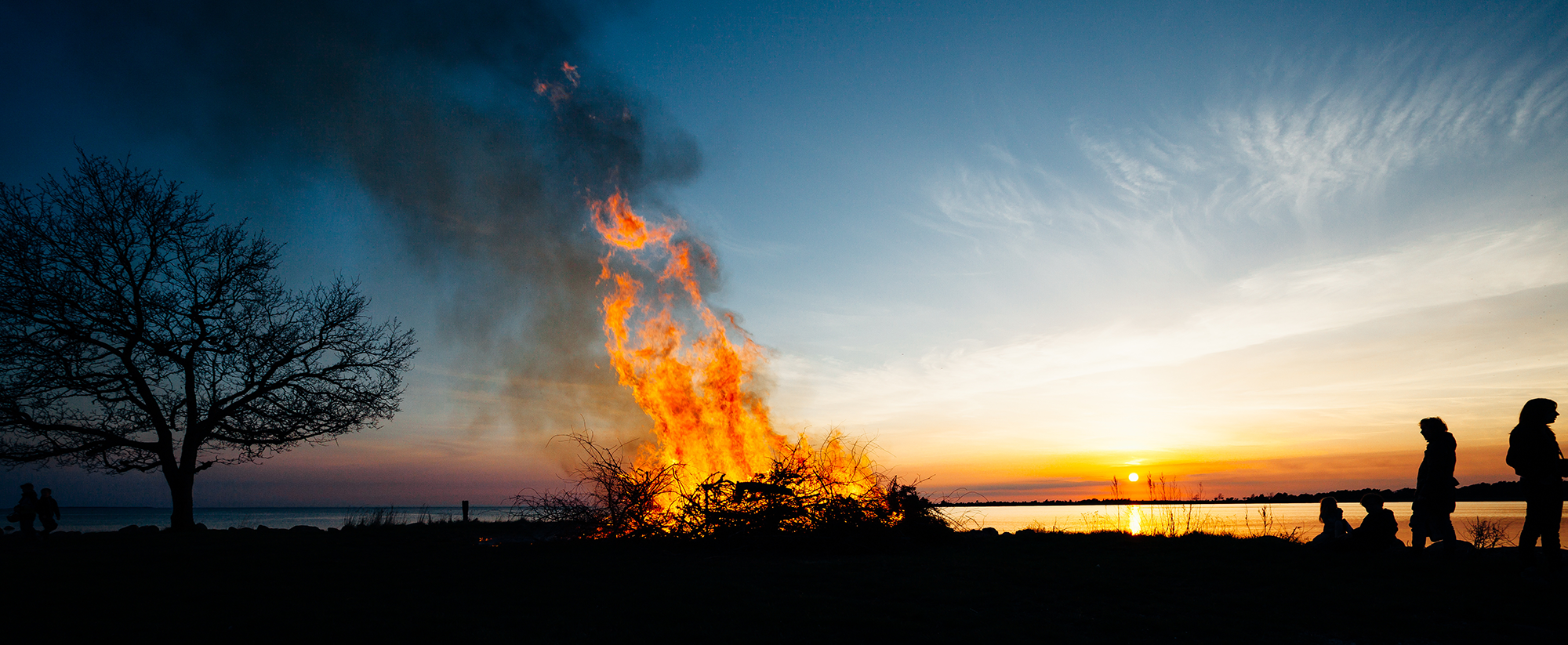 Bilden visar en större brasa som brinner vid en strandkant. Det är solnedgång.
